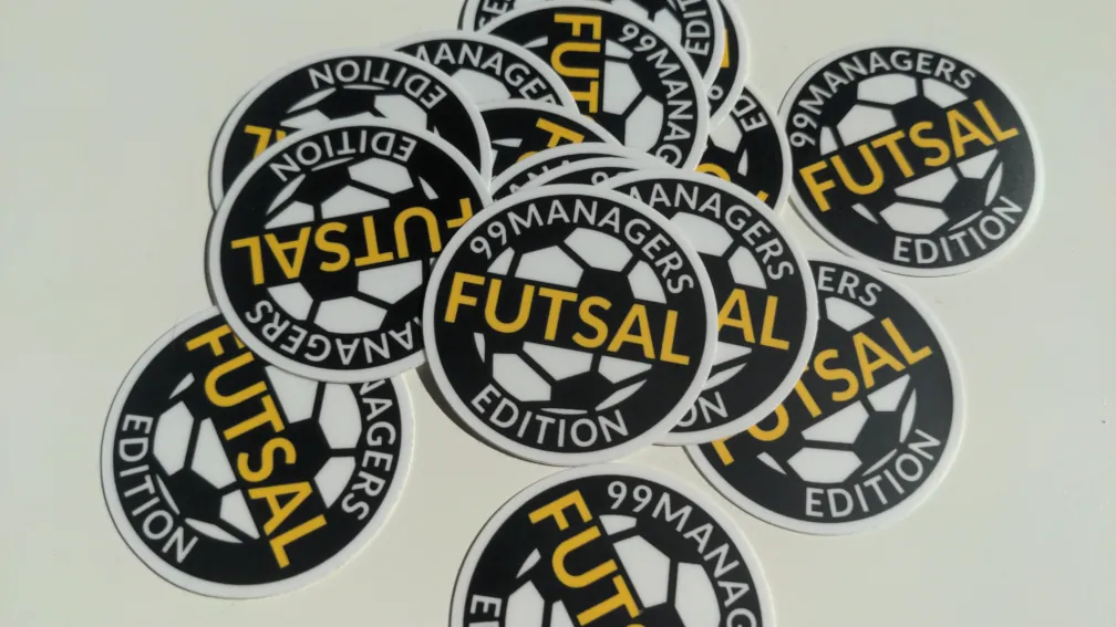 Picuter of stickers spread across the table showing the logo with the text '99MANAGERS FUTSAL EDITION'
